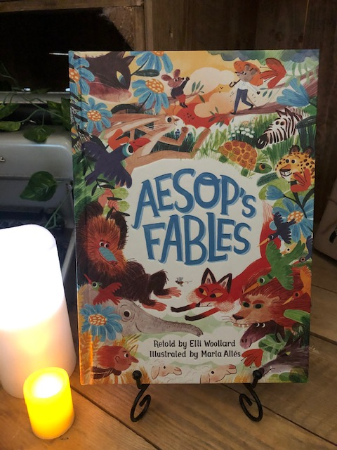 Image of the front cover of the hardback book of Aesop's Fables stood in a book stand with candles
