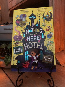 Image of the front cover of the paperback book The Nothing to See Here Hotel, written by Steven Butler and illustrated by Steven Lenton. Displayed on a book stand.