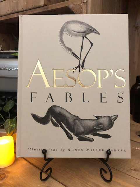 Image of the front cover of the cloth bound hardback book of Aesop's Fables in a book stand with candles