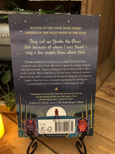 Image of the back cover of the paperback book The Girl Who Speaks Bear, written by Sophie Anderson and illustrated by Kathrin Honesta. Displayed on a book stand with candles.