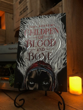 Load image into Gallery viewer, Image of front cover of paperback book Children of Blood and Bone stood in book stand with a candle