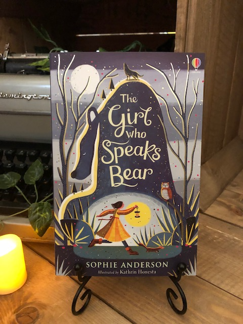Image of the front cover of the paperback book The Girl Who Speaks Bear, written by Sophie Anderson and illustrated by Kathrin Honesta. Displayed on a book stand with candles.