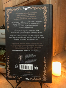 Image of the back cover of the hardback book The Sisters Grimm, written by Menna van Praag. Displayed on a book stand with candles.