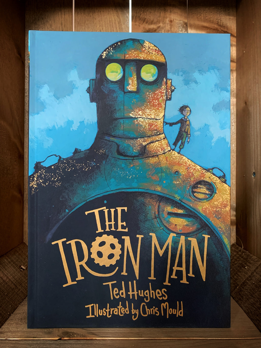 Image showing the front cover of The Iron Man. The cover features an illustration of a close up of the iron mans head and shoulders, with Hogarth standing on his shoulder, and a blue sky behind. The title is written at the bottom in foiled gold lettering, and gold is scattered across the illustration.