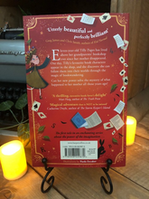 Load image into Gallery viewer, Image of the back of the paperback book Pages &amp; Co: Tilly and the Bookwanderers written by Anna James and illustrated by Paola Escobar. Displayed on a book stand with candles.