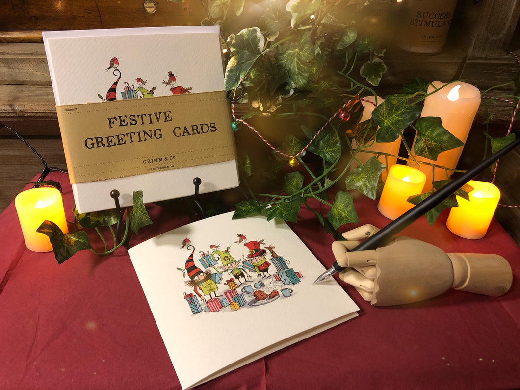 Image of a pack of 5 greetings cards showing a cartoon illustration of three shop elves amid present wrapping and snacks drawn by Chris Mould. Display shows a wooden mannequin hand holding a Dip Wand poised to write a card