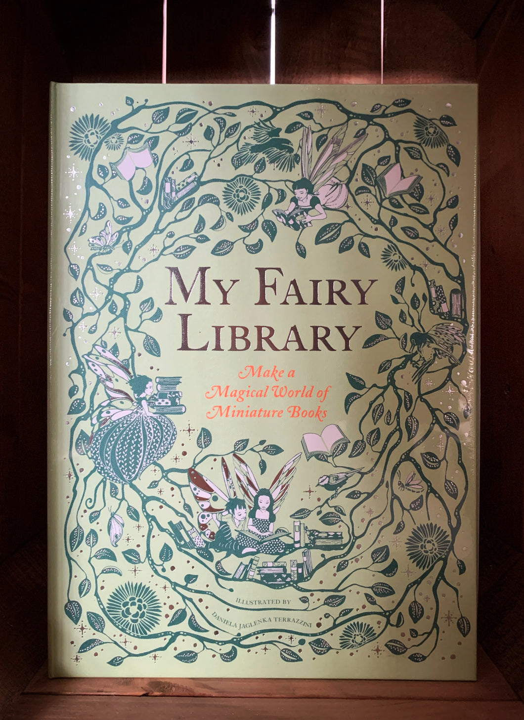 Image of the front of the box for My Fairy Library. The front has a pale green background, with the name in the center in foiled gold lettering. Surrounding the title are illustrations of leaved vines and flowers in dark green, with fairies and books scattered throughout in dark green and white. 