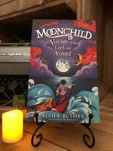 Image of the front of the paperback book Moonchild Voyage of the Lost and Found by Aisha Bushby and illustrated by Rachael Dean. Displayed on a book stand with candles.