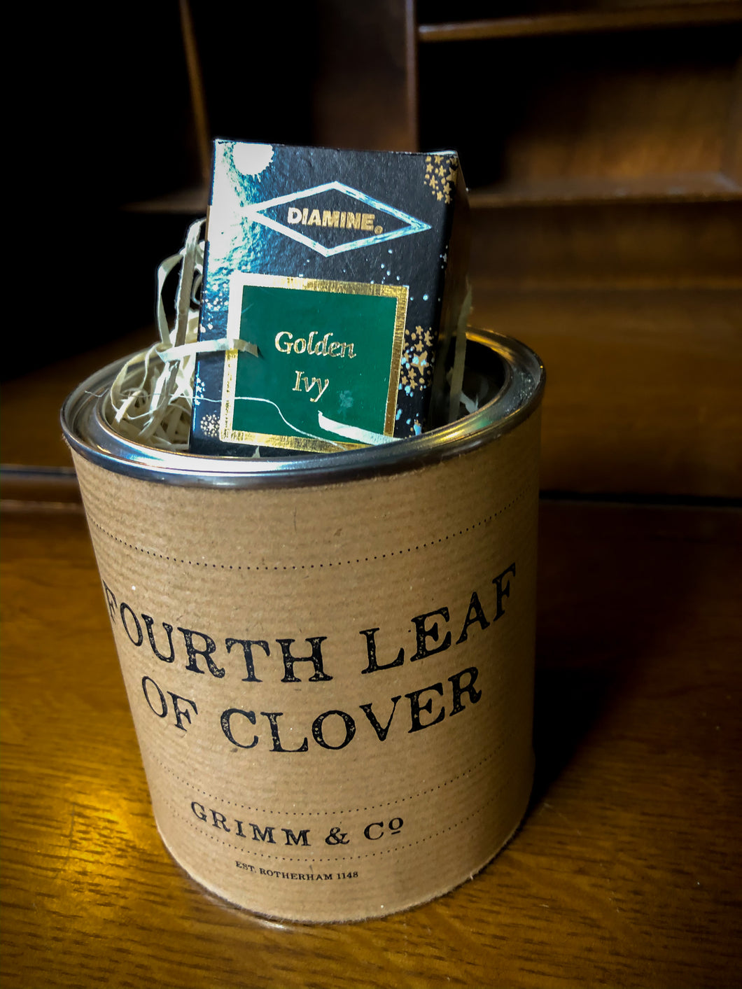 Image of a tin of Fourth Leaf of Clover with kraft paper label and the box of ink poking out of the tin among wood wool shreds