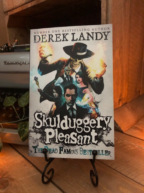Image of the front of the paperback book Skulduggery Pleasant by Derek Landy. Displayed on a book stand.