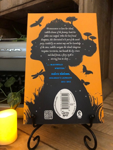 Load image into Gallery viewer, Image of the back cover of the paperback book The Girl of Ink and Stars, written by Kiran Millwood Hargrave. Displayed on a book stand with candles.