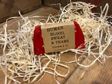 Load image into Gallery viewer, Image of Human Blood, Sweat &amp; Tears bar, a red pomegranate scented soap slice shown with a kraft paper label. Soap contains oats which can be seen throughout the slice of soap.