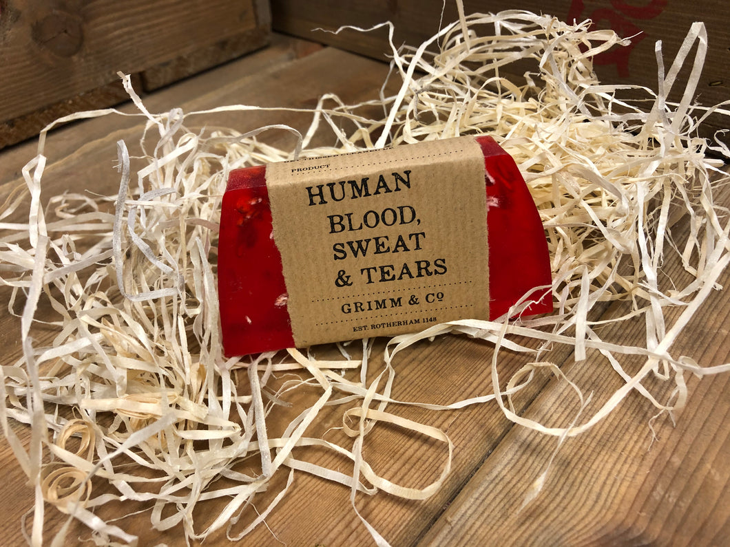 Image of Human Blood, Sweat & Tears bar, a red pomegranate scented soap slice shown with a kraft paper label. Soap contains oats which can be seen throughout the slice of soap.