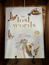 Load image into Gallery viewer, Image of the front of the book The Lost Words. The cover has a plain white/cream background, with illustrations of 5 goldfinches in flight, and a dandelion seed head. The title of the book is written in the center in foiled gold text.