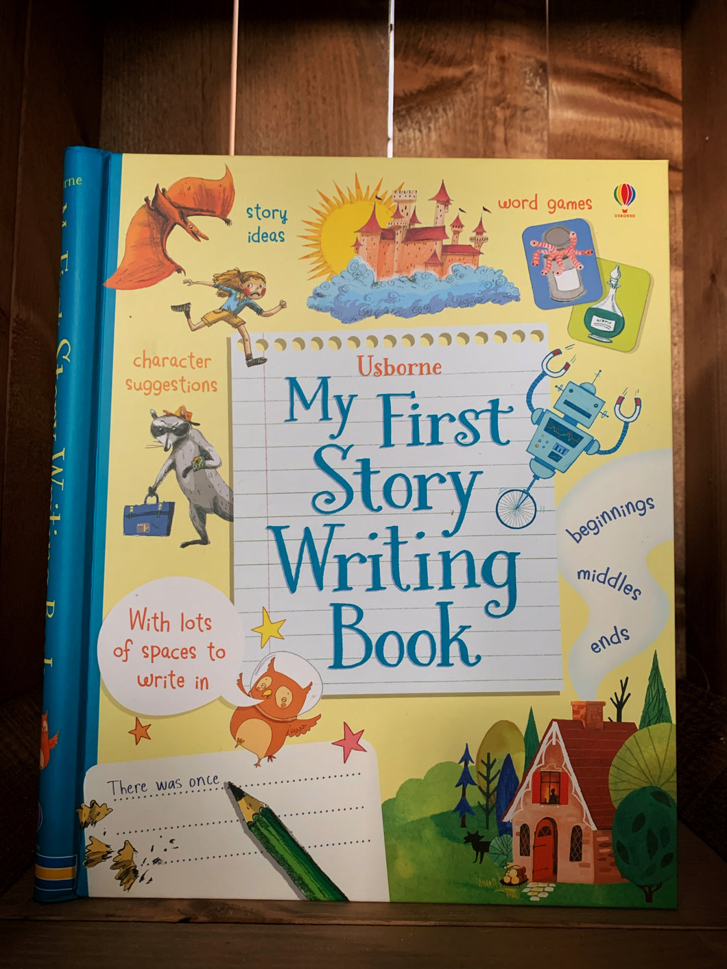 Image of the front cover of My First Story Writing Book. The cover has a yellow background with a blue spine, and features various settings and characters that could feature in stories. This includes a house in a forest, a castle in the clouds, a robot, a raccoon holding a suitcase, and a dragon chasing someone. It also features text explaining what kinds of  tips the book has inside. 