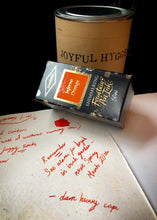 Load image into Gallery viewer, Image of the Joyful Hygge ink in box next to tin, with a parchment page of writing done in the orange and gold shimmering ink.