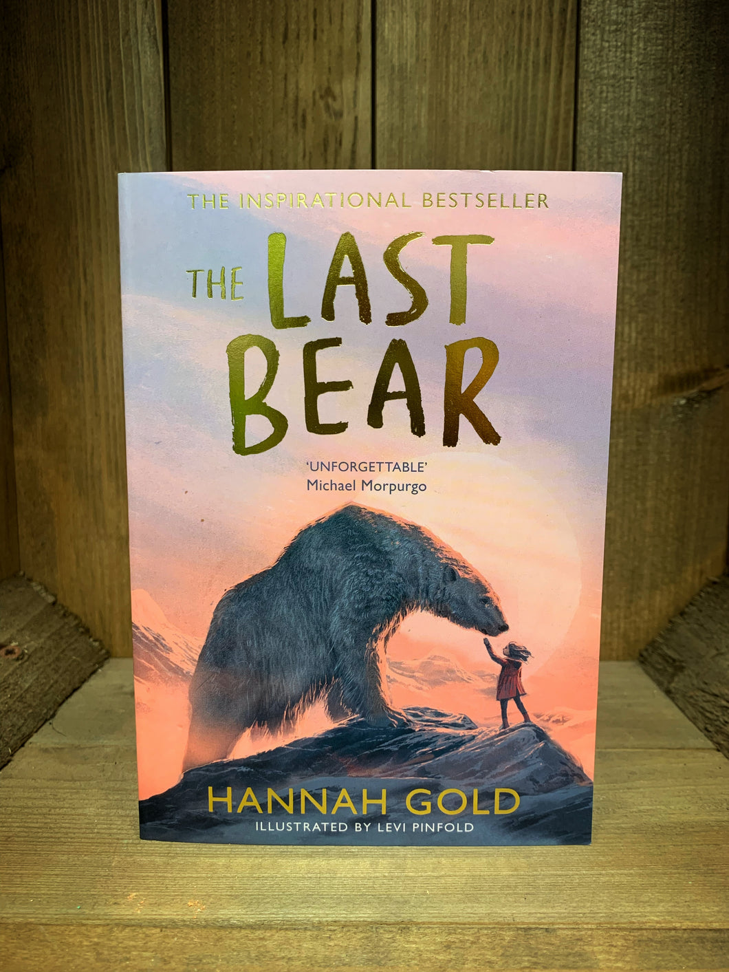 Image of the front cover of the book The Last Bear. The cover shows an illustration of a girl and a polar bear standing on a mountaintop in front of a sunrise.
