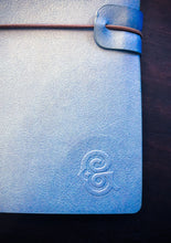 Load image into Gallery viewer, Close up image of the embossed Grimm &amp; Co monogram on the bottom right corner of a blue vegan leather journal.