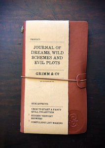 Top down image of a Burgundy coloured Journal of Dreams, Wild Schemes, and Evil Plots. Otherwise known as a vegan leather, refillable journal with a matching coloured elasticated closure around the middle. It is embossed with the Grimm & Co monogram in the lower right corner, and has a brown kraft label wrapped around two thirds of the cover. 