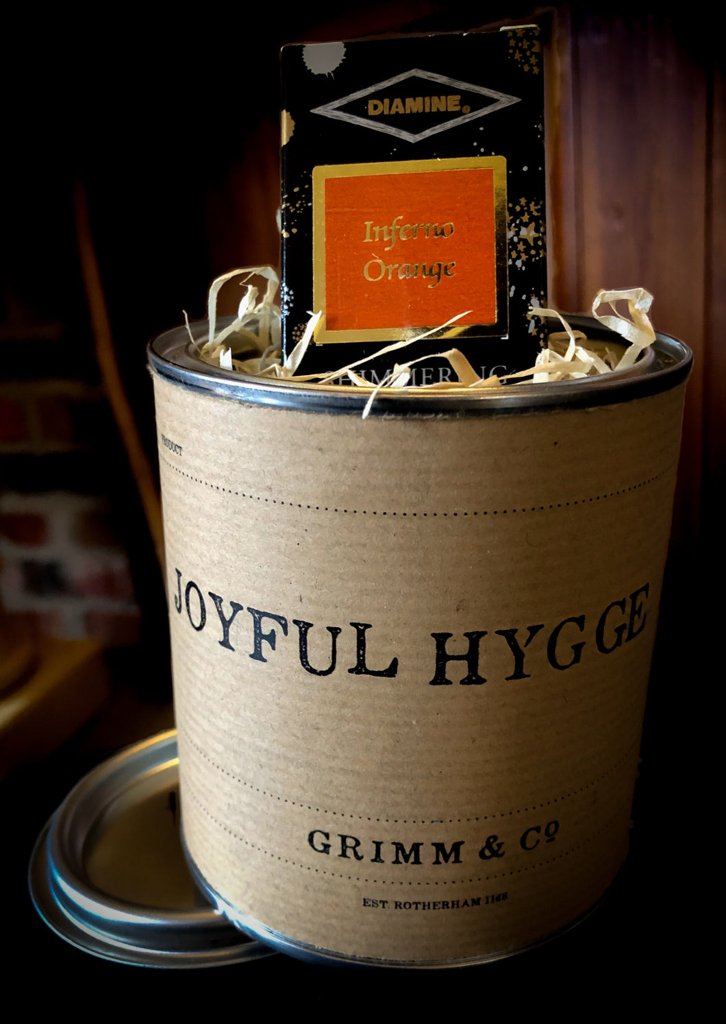 Joyful Hygge, a tin containing orange and gold shimmering ink suitable for writing and drawing. Ink box is shown poking out of tin filled with wood wool.