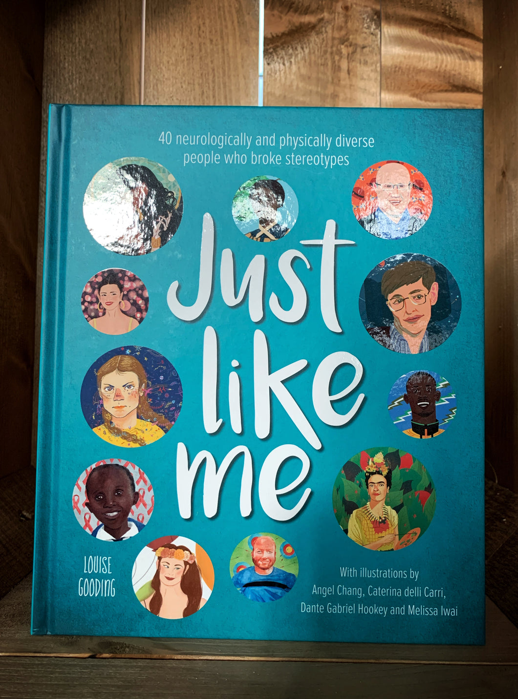 Image of the front cover of the hardback book Just Like Me. The cover has 11 round illustrated portraits of people featured within the book, set on a dark teal background.