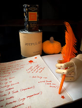 Load image into Gallery viewer, Image of the Joyful Hygge ink in box next to tin, with a parchment page of writing done in the orange and gold shimmering ink and a wooden mannequin hand holding an orange feather quill.