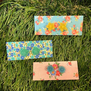 Image shows three designs of earrings yellow daffodils, aqua roses and blue daisies all made from acrylic shown displayed on fake grass.