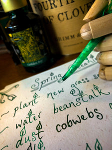 Image of Fourth Leaf of Clover green and gold shimmer ink displayed with a wooden mannequin gand and feather quill poised over paper with green and gold shimmer cursive writing