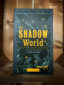 Image of the front of the box for The Shadow World: Storytelling Card Game. The background is dark grey, with an illustration of a factory in black and light grey, with cogs, wheels, pipes, and stairs leading up to a lit up town on a platform at the top of the box, with various animals, robots, fish and individuals scattered throughout. The title is in the center in bright yellow text. 