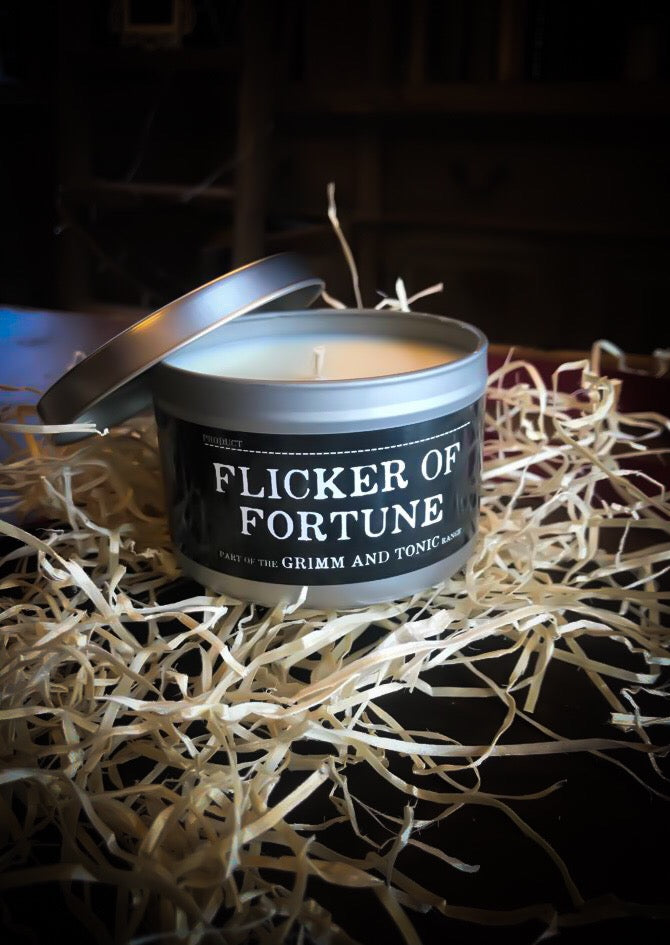Image of a candle in a tin called Flicker of Fortune, lid is removed to reveal candle wax and wick inside