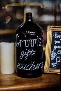 Image of a brown glass bottle with the words Grimm's Gift Vouchers written on in white chalk pen. Bottle is on a counter with a candle.