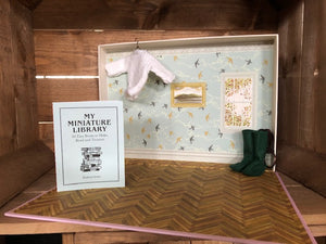 Image showing the inside of the box for My Miniature Library and the instruction booklet that comes with the kit. The inside of the box is decorated to look like a library space. The image also includes a fairy jumper hanging from the box and some pixie boots, which are not included within the kit. 