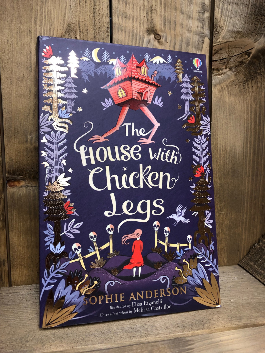 Image of the paperback book The House with chicken Legs with a purple cover illustrated with a border of trees and a girl standing by a fence with skulls on the top and a house with chicken legs at the top of the cover.