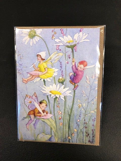Image shows the Fairy Land greetings card with daisies. Three fairies sit on a daisy plant writing letters on leaves with daisy petal quills. Card is in full-colour with a pale blye background and other wild flowers in the scene. Card comes with a kraft envelope.