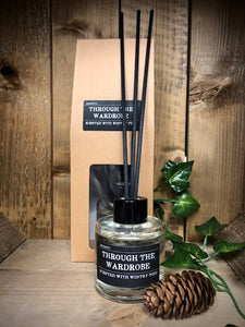 Image shows the Through the Wardrobe Scent Portal reed diffuser set with the kraft packaging and 100ml glass bottle with black lid and reeds, displayed with a  pine cone and some evergreen ivy.