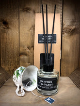Load image into Gallery viewer, Image shows the Hatter&#39;s Tea Party Scent Portal reed diffuser set with the kraft packaging and 100ml glass bottle with black lid and reeds, displayed with a patterned china tea cup and an Earl Grey tea bag.