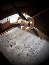 Load image into Gallery viewer, Image of a matte black Dip Wand in a wooden mannequin hand poised above paper with cursive writing. Dip Wand is a wooden dip pen with nib for use with writing or drawing inks