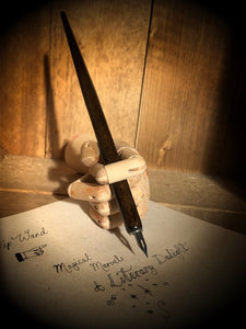 Image of a Dip Wand in a wooden mannequin hand poised above paper with cursive writing. Dip Wand is a wooden dip pen with nib for use with writing or drawing inks