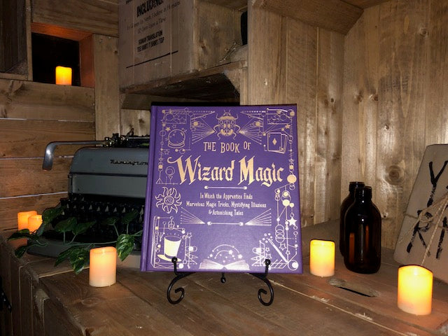 Image of the front cover for The Book Of Wizard Magic, written by Janice Eaton Kilby. The lighting of the image shows off the reflective lettering on the cover. Displayed on a book stand with candles..