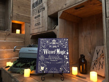 Load image into Gallery viewer, A further back image showing the front cover for The Book Of Wizard Magic, written by Janice Eaton Kilby. Displayed on a book stand with candles.