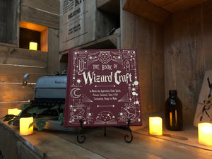 Image shows the front cover of The Book Of Wizard Craft, written by Janice Eaton Kilby. Displayed on a book stand with candles.
