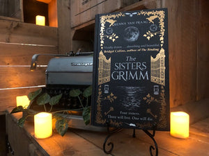 Image of the front cover of the hardback book The Sisters Grimm, written by Menna van Praag. Displayed on a book stand with candles.