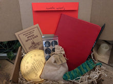 Load image into Gallery viewer, Image of the Gift Box to Mull Over containing a soft touch red faux leather notebook with pencil, a set of 12 Grimm &amp; Co stickers, an envelope of writing prompts, spiced apple juice sachet, a giant chocolate coin, a knitted elf stocking decoration and a jute drawstring pouch of edible elf staffs, all presented in a kraft cardboard box filled with wood wool.