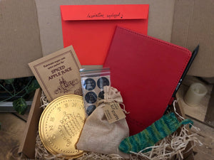 Image of the Gift Box to Mull Over containing a soft touch red faux leather notebook with pencil, a set of 12 Grimm & Co stickers, an envelope of writing prompts, spiced apple juice sachet, a giant chocolate coin, a knitted elf stocking decoration and a jute drawstring pouch of edible elf staffs, all presented in a kraft cardboard box filled with wood wool.