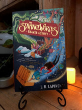 Load image into Gallery viewer, Image of the front cover of the paperback book The Strangeworlds Travel Agency, written by L.D. Lapinski. Displayed on a book stand with candles.