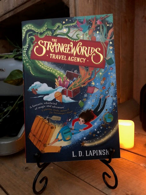 Image of the front cover of the paperback book The Strangeworlds Travel Agency, written by L.D. Lapinski. Displayed on a book stand with candles.