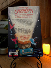 Load image into Gallery viewer, Image of the back cover of the paperback book The Strangeworlds Travel Agency, written by L.D. Lapinski. Displayed on a book stand with candles.