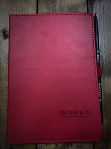 Image of the red soft touch faux leather notebook and Word Wand pencil as included in the Gift Box to Mull Over. Notebook is embossed in the bottom right corner with Grimm & Co Apothecary to the Magical