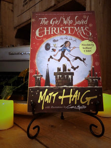 Image of the front cover of the paperback book The Girl Who Saved Christmas, written by Matt Haig and illustrated by Chris Mould. Displayed on a book stand with candles.