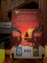 Load image into Gallery viewer, Image of the back cover of the paperback book The Girl Who Saved Christmas, written by Matt Haig and illustrated by Chris Mould. Displayed on a book stand with candles.